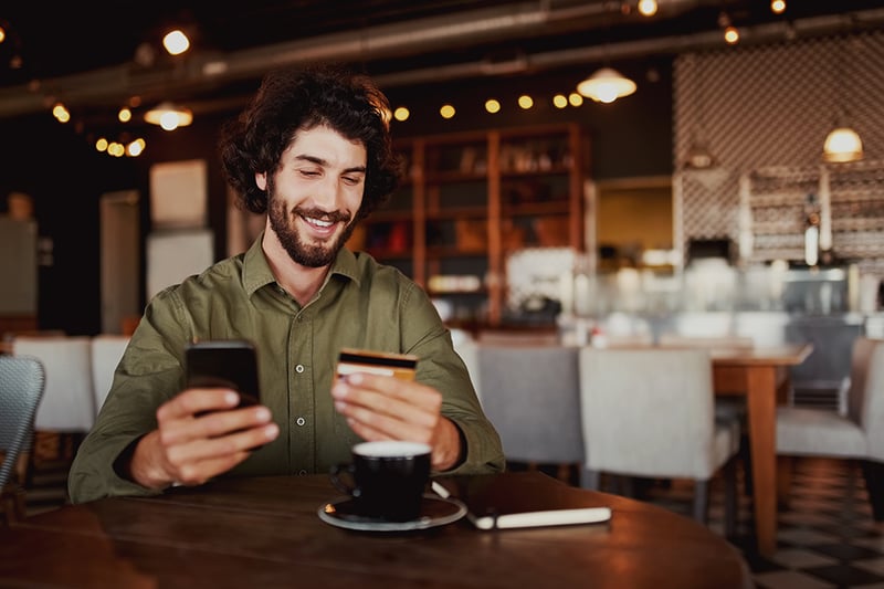 Man in a coffee shop holding a cell phone and a credit card.