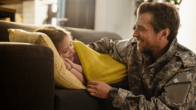 United States service member laughing with their child and tucking them in on the couch.