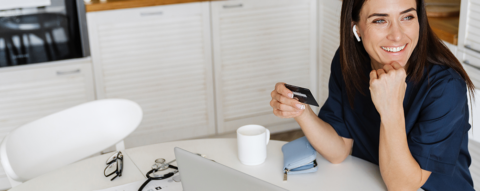 Wide image of a person sitting at their desk, holding their credit card and looking out the window smiling