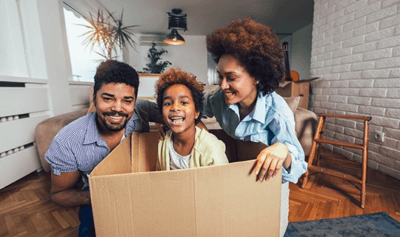 A three person family in a living room smiling, with the child inside a cardboard box in the center