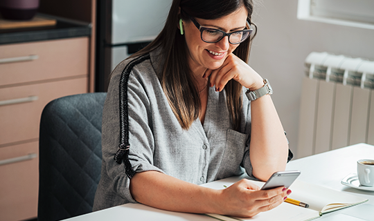 A smiling person at desk, wearing wireless earbuds looking down at their mobile phone