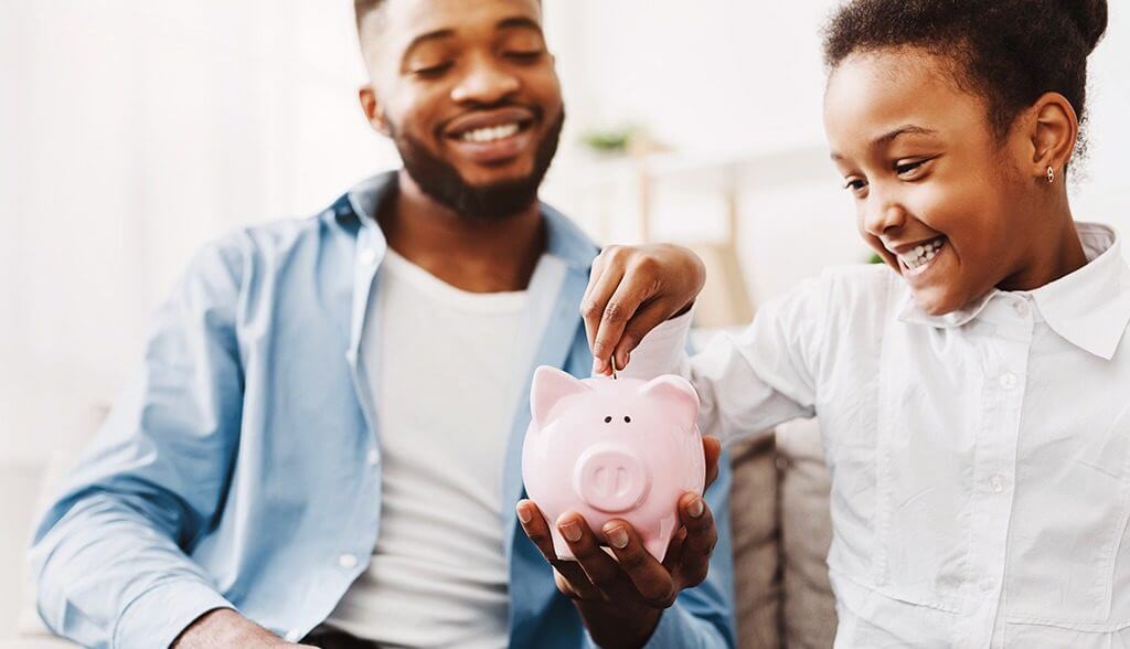 Little girl putting money in piggy jar being held by her dad