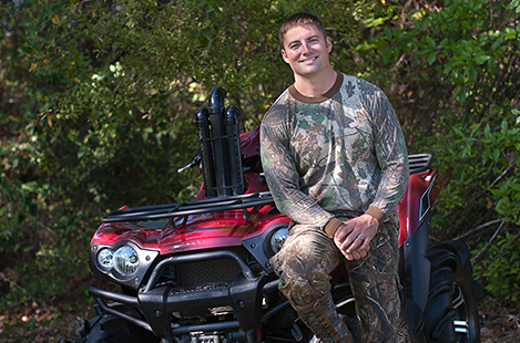 A person wearing camouflage sitting on their motorcycle with foliage in the background