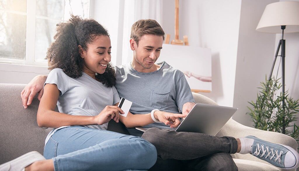 young couple sitting on couch, using credit card on laptop